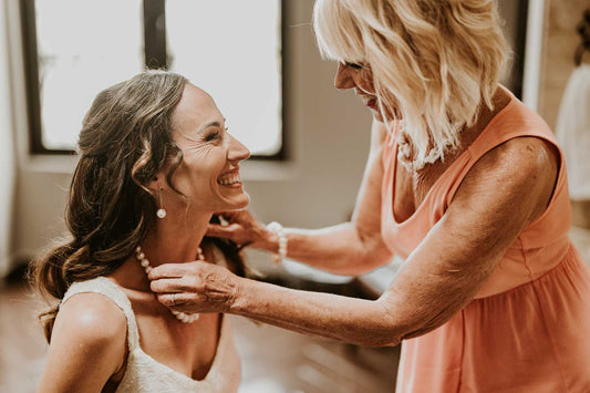 A joyful moment between two women as one, dressed in a sleeveless wedding gown, smiles while the other, wearing a sleeveless peach-colored dress, places a pearl necklace around her neck. The bride also wears matching pearl earrings. The intimate indoor se