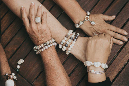 Three women’s arms shown adorned with various freshwater pearl bracelets and rings. The bracelets feature large, irregularly shaped pearls in shades of white and light purple, with silver and gold accents. The image is set on a dark wooden table, highligh