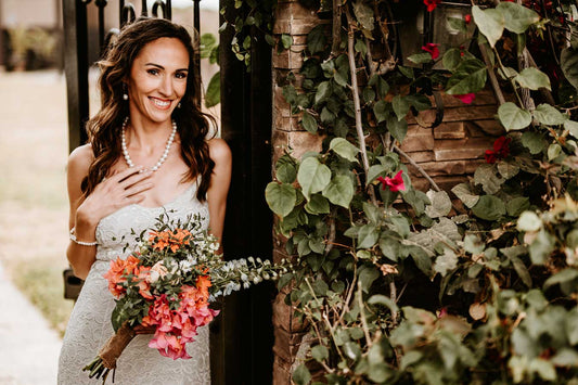 A bride wearing a pearl necklace and matching earrings smiles gently with her eyes closed, her elegant white gown glowing softly in a low-lit room.